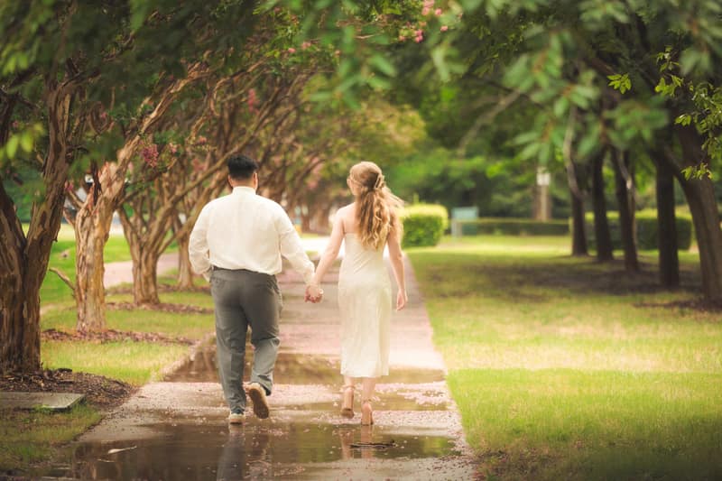 Couple walking hand in hand away from the camera down a rain-wet path under arching crepe myrtle trees, pink petals floating in a puddle