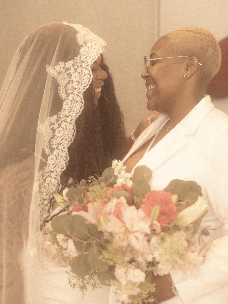 Two brides laughing face to face on their wedding day, one under a scalloped lace veil, a pink and coral bouquet between them