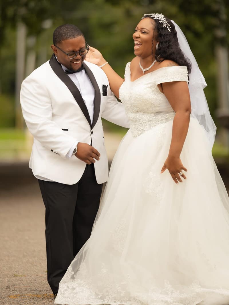 Bride throwing her head back in laughter as she touches the groom's cheek on a leafy lane, both dressed in white