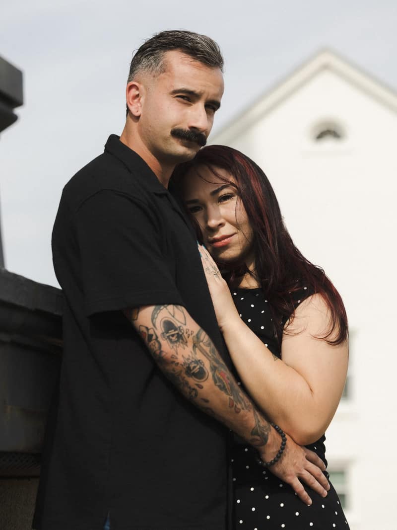 Tattooed man in black holding his partner close as both look into the camera in front of a white clapboard gable