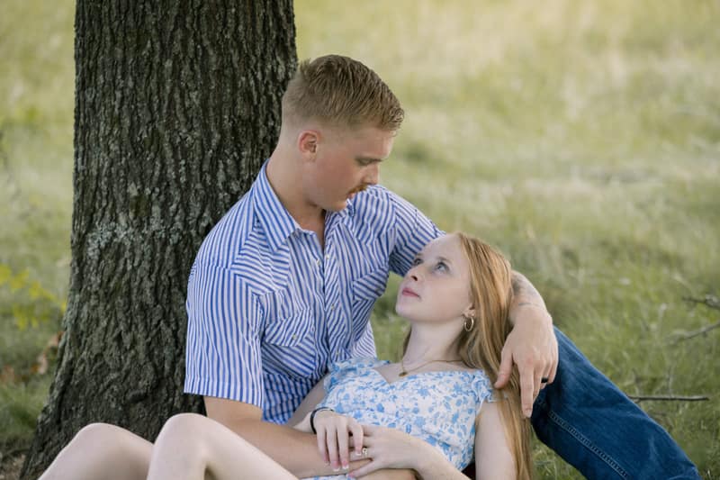 Woman lying back in her partner's lap beneath an oak tree, gazing up at him as he looks down at her