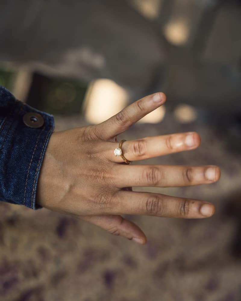 Hand held out to show a new gold solitaire engagement ring, denim cuff at the wrist and warm bokeh behind