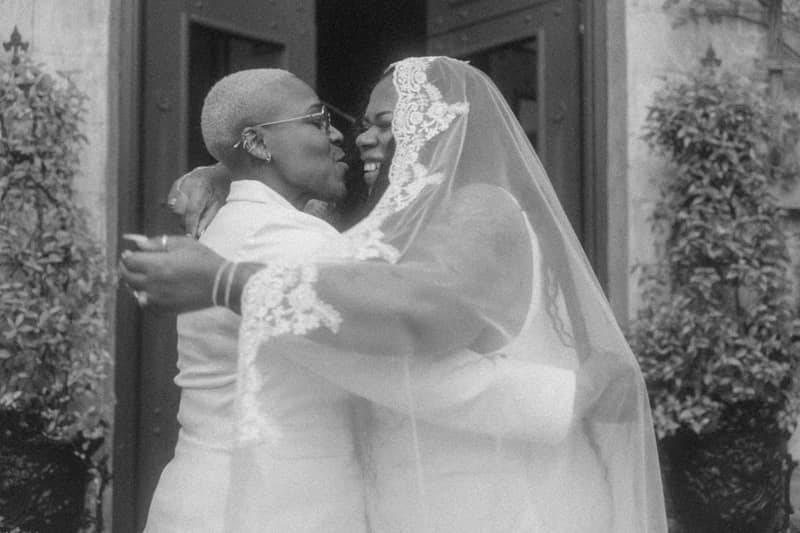Newlyweds embracing in black and white before dark wooden doors, one kissing the laughing bride's cheek through her lace veil