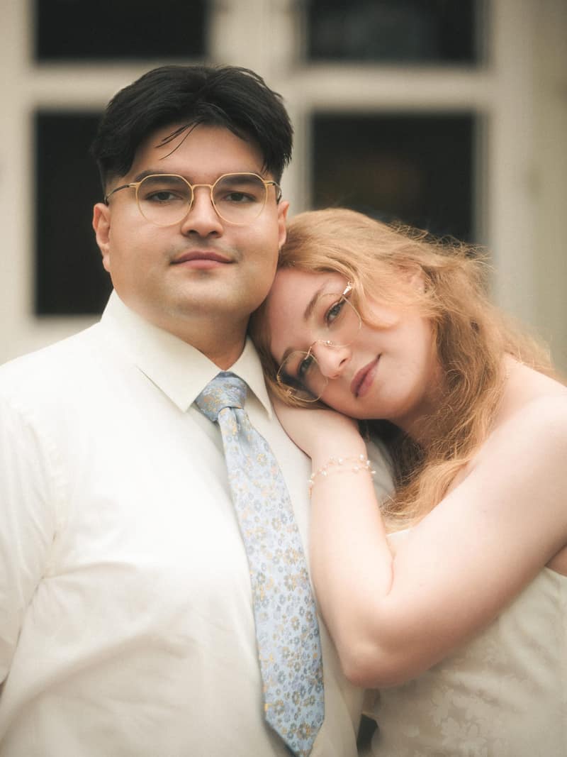 Engaged couple close together, her head resting on his shoulder as both look softly into the camera in front of a pale doorway