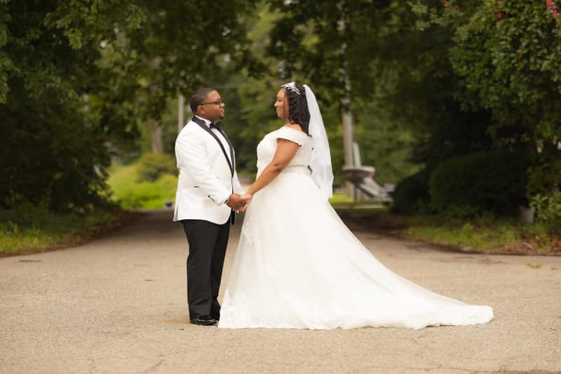 Bride and groom holding hands and gazing at each other under a tree canopy, her long train spread across the road