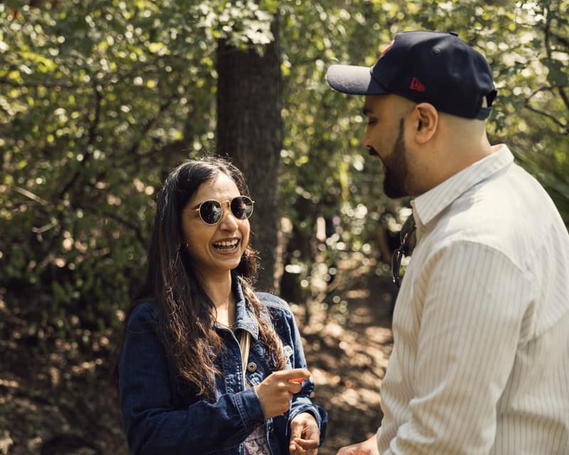 Woman in round sunglasses laughing just after saying yes as her partner grins down at her in a sunlit forest