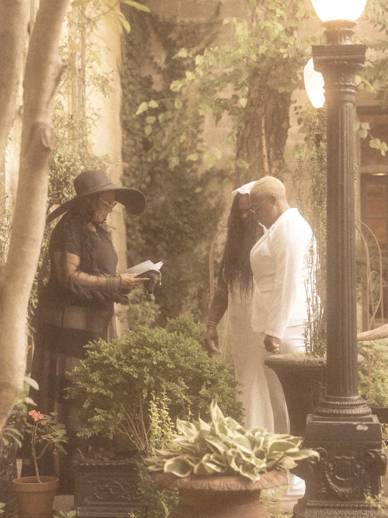 Garden wedding ceremony seen through foliage, the officiant reading as the couple stands with heads bowed beside an iron lamp post