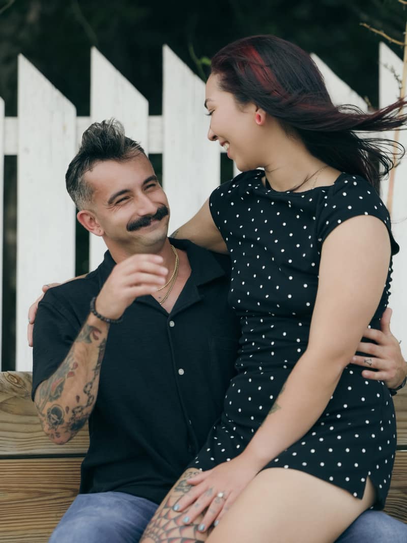 Couple laughing together on a wooden bench in front of a white picket fence, her hair caught in the wind