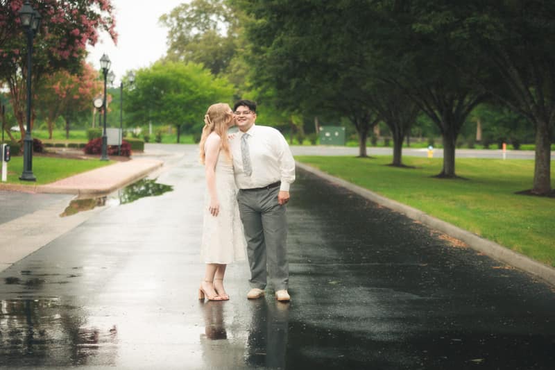 Woman kissing her partner's cheek as he grins, standing in the middle of a glossy rain-soaked lane lined with trees