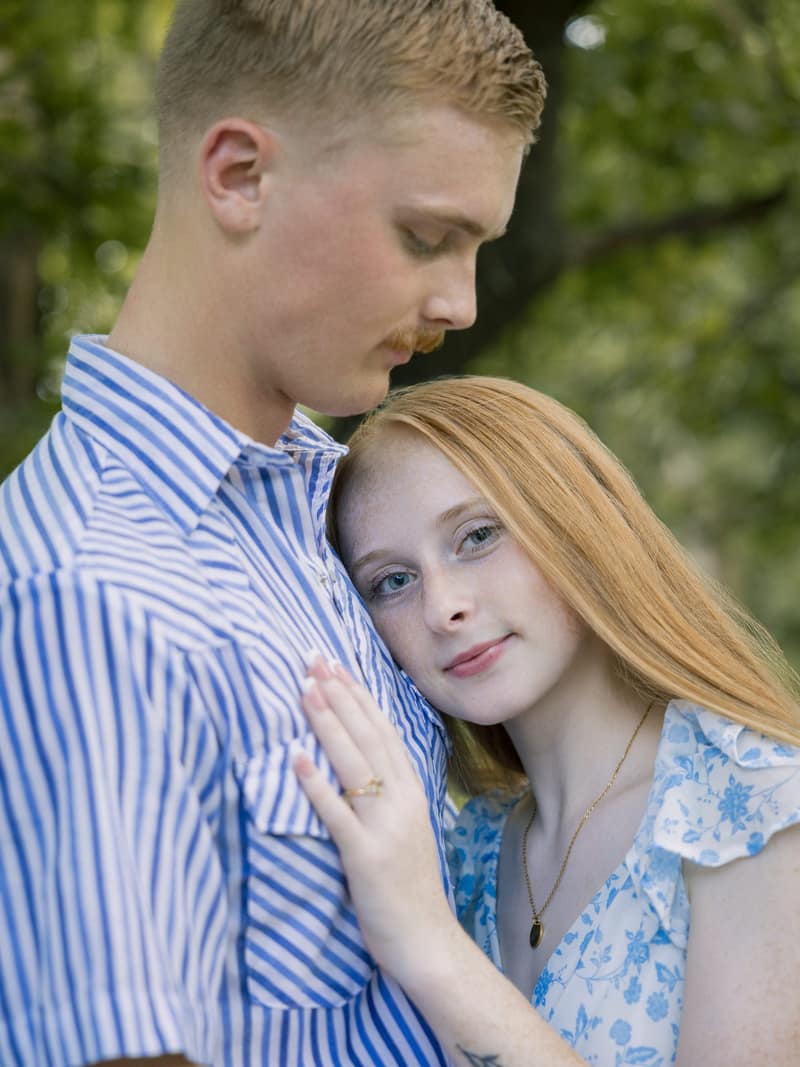 Freckled redhead resting her head on her partner's chest and looking into the camera, her engagement ring on the hand at his shirt