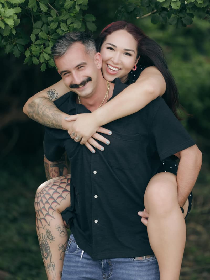Woman riding piggyback on her partner, both grinning at the camera beneath dark green leaves