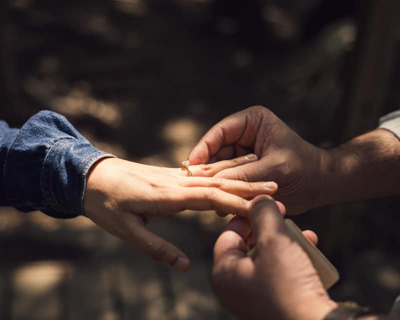 Hands slipping a gold solitaire ring onto her finger in dappled forest shade, a denim cuff at the edge of the frame
