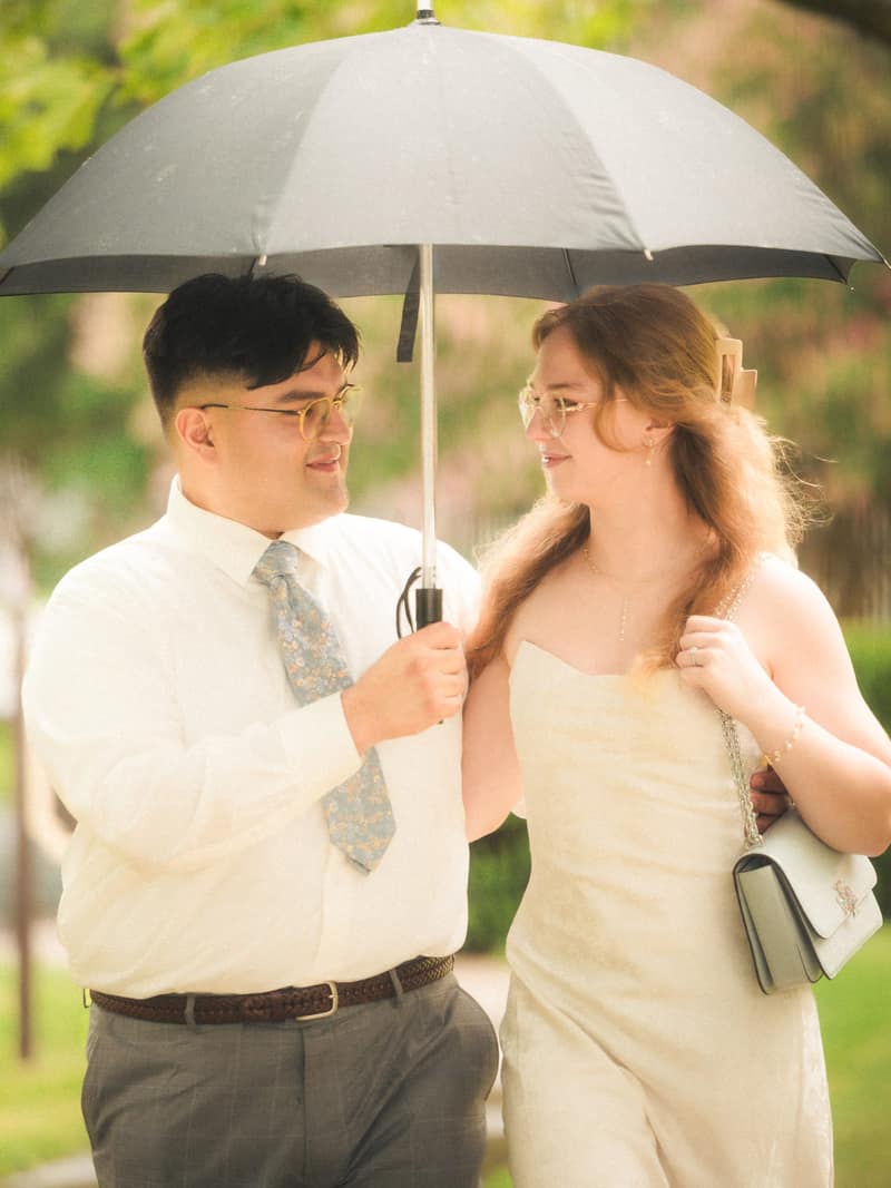 Couple sharing a grey umbrella and smiling at each other as they walk through a glowing green park after the rain
