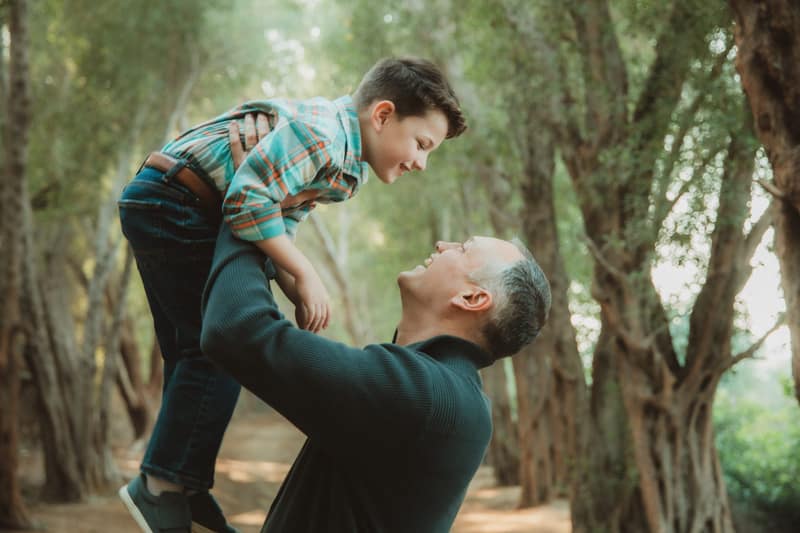 Father lifting his laughing son overhead, the two grinning at each other nose to nose in a sun-dappled tunnel of trees