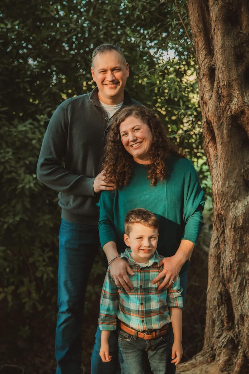 Mother, father and young son posed together beside a large tree trunk, all smiling at the camera in warm light