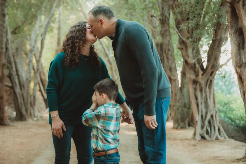 Parents sharing a kiss on a woodland path while their young son covers his eyes between them