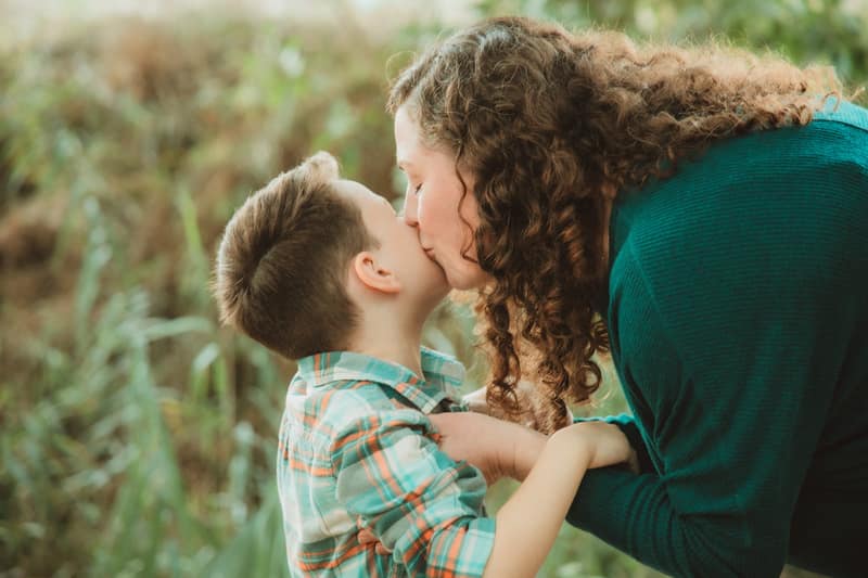 Mother leaning down to kiss her son's cheek, her curls framing the moment against soft golden grasses