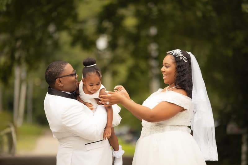 Groom holding a toddler in a white dress on the wedding day as the smiling bride reaches out to take her tiny hands