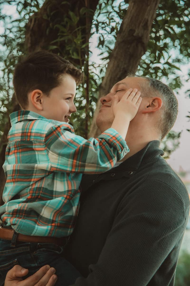 Young boy in his father's arms cupping his dad's face and smiling up at him, the dad's eyes closed in a grin under a leafy tree