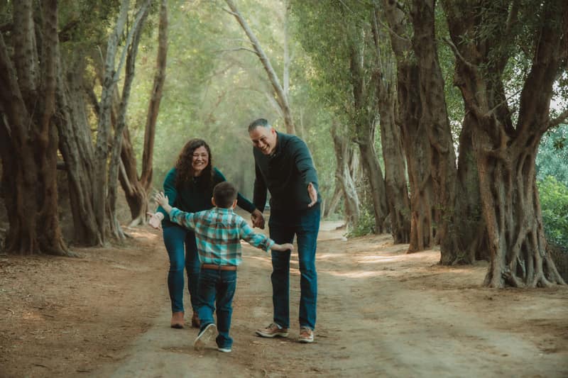 Young boy running with arms wide toward his laughing parents down a misty tree-lined dirt path