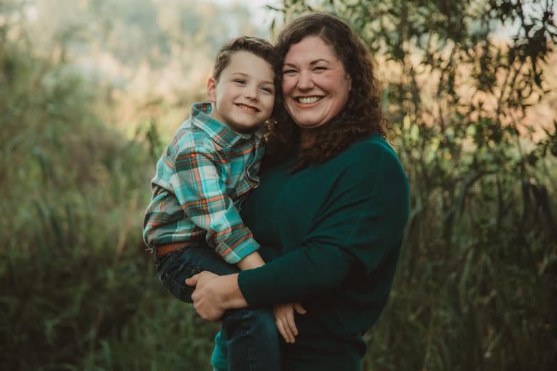 Mother holding her son cheek to cheek, both beaming at the camera against warm backlit reeds