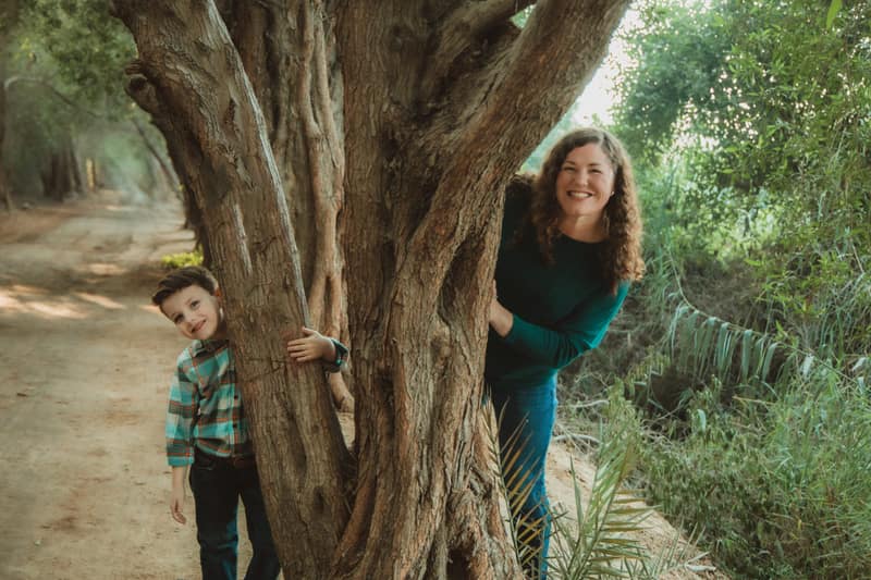 Mother and son playfully peeking out from either side of a massive twisted tree trunk beside a dirt path