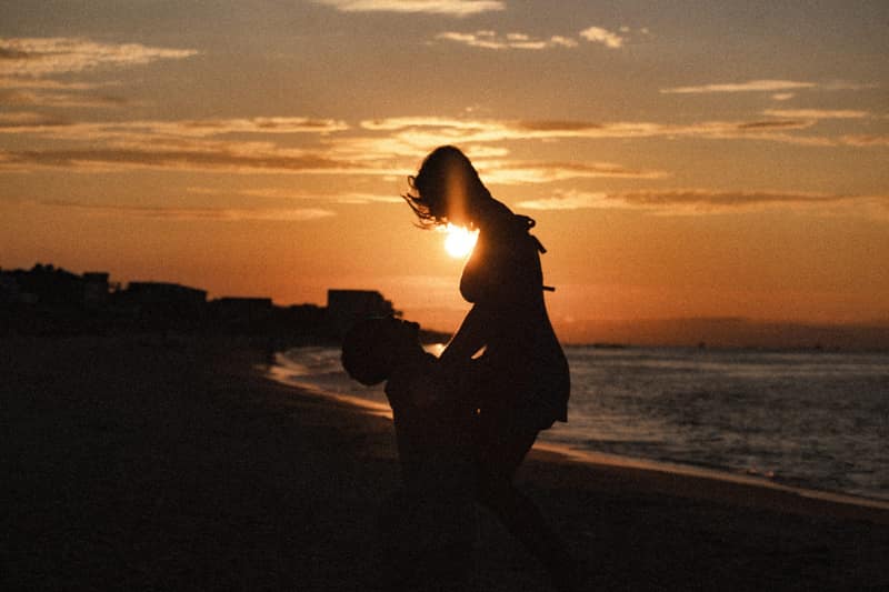 A man lifts his partner into the air on an empty beach at sunset, their silhouettes framed against a glowing sky as the sun breaks through between them