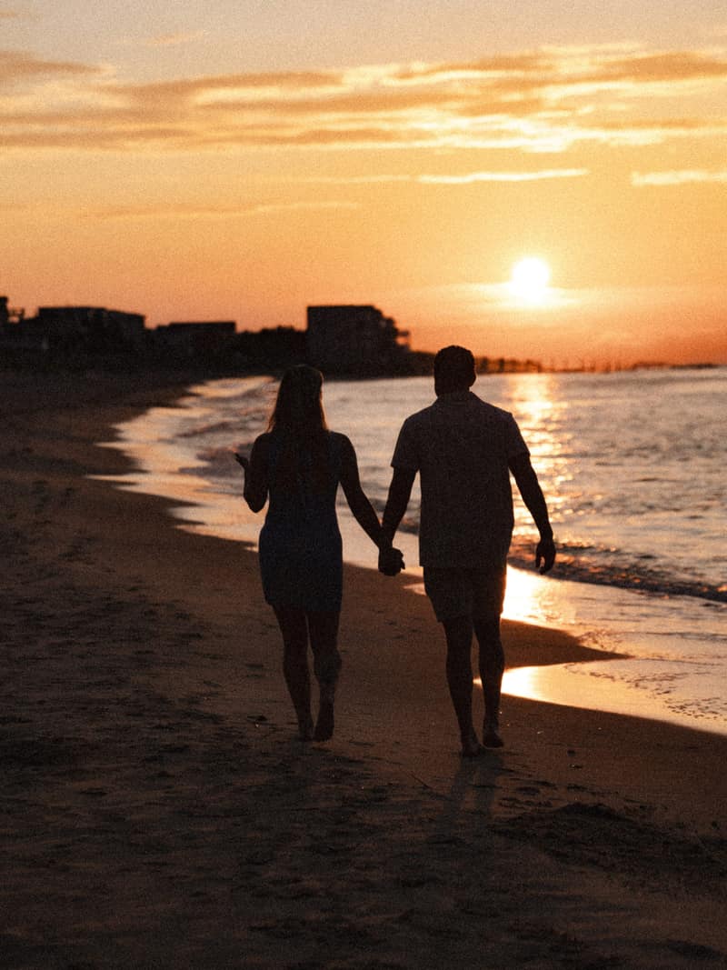 A couple walks barefoot hand in hand along the wet shoreline at sunset, silhouetted against a gold sky reflected in the water