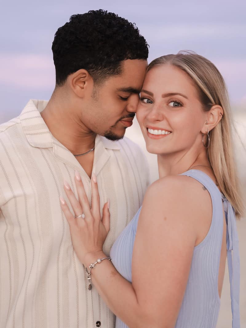 A couple stands cheek to cheek at dusk on the beach, her engagement ring resting on his chest as she smiles toward the camera