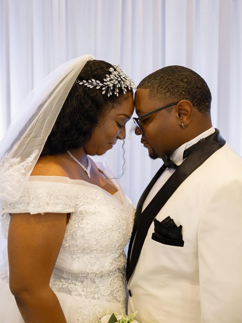 A bride and groom rest their foreheads together with eyes closed in front of softly lit white ceremony drapery