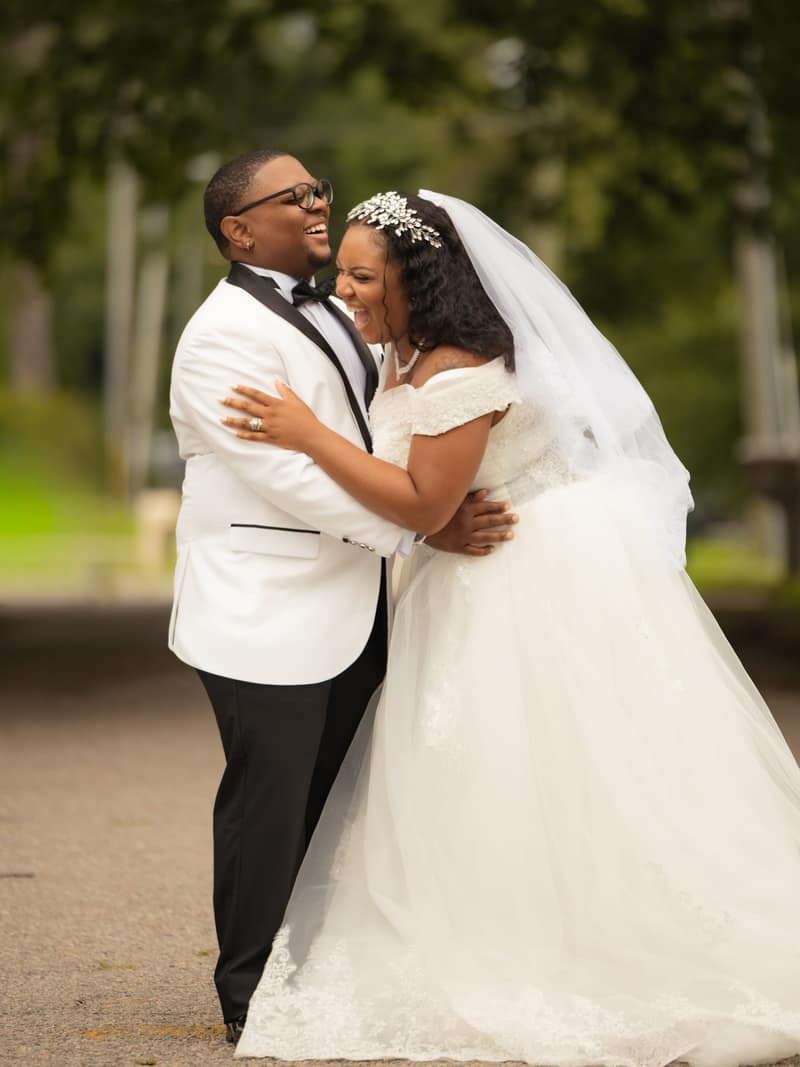 A bride and groom laugh together in a close embrace on a tree-lined path, her veil and lace gown catching soft overcast light