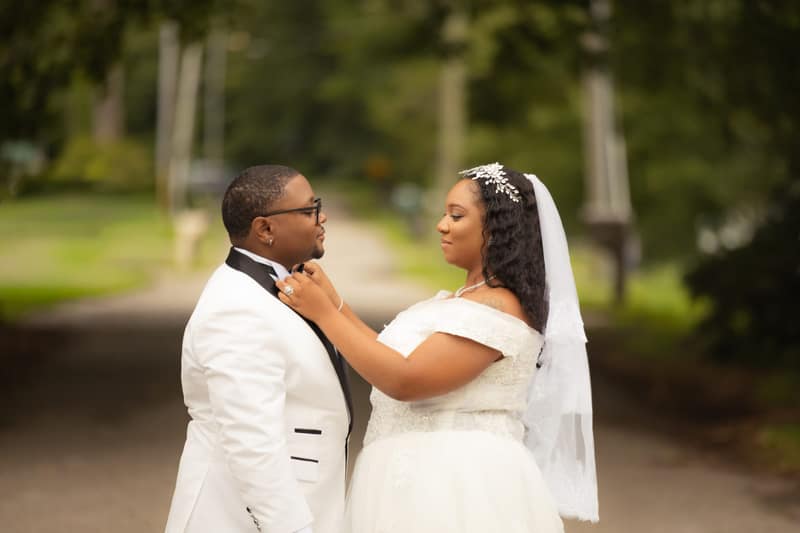 A bride straightens her groom's bow tie as the two look at each other on a quiet park path framed by green trees