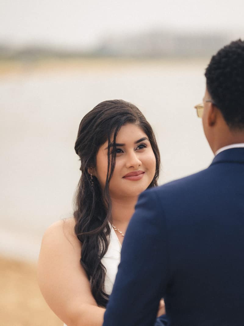A bride smiles softly at her groom over his shoulder, her dark hair loose in the wind against a pale blurred seascape