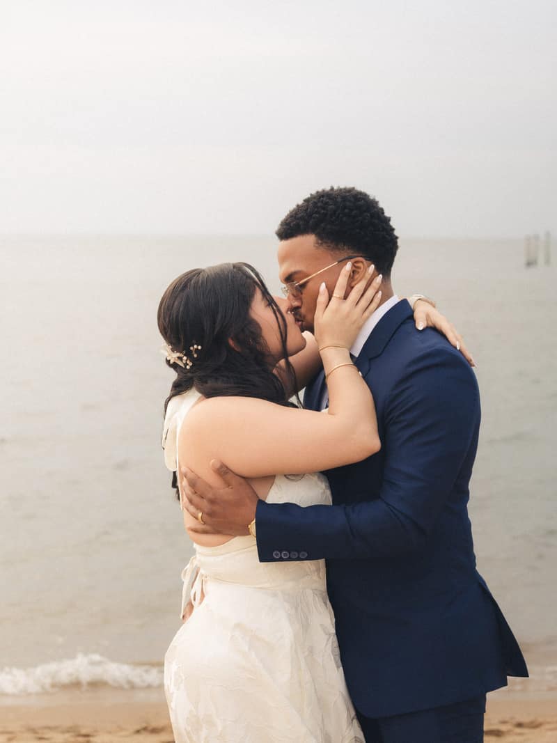 A bride and groom kiss on a foggy beach, her hand cradling his face and his arm around her waist, pale water and sky behind them