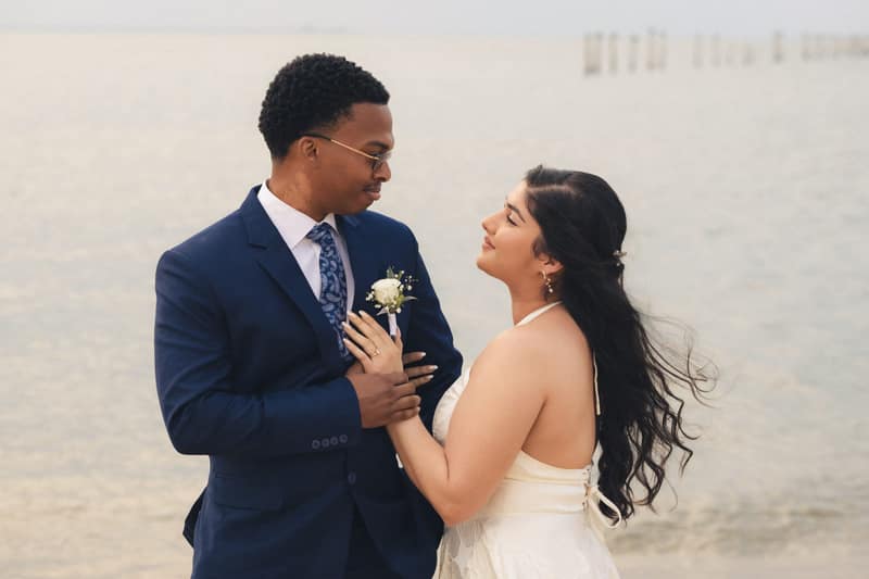 A bride in an ivory dress rests her hand on her groom's navy lapel as they look into each other's eyes on a hazy shoreline